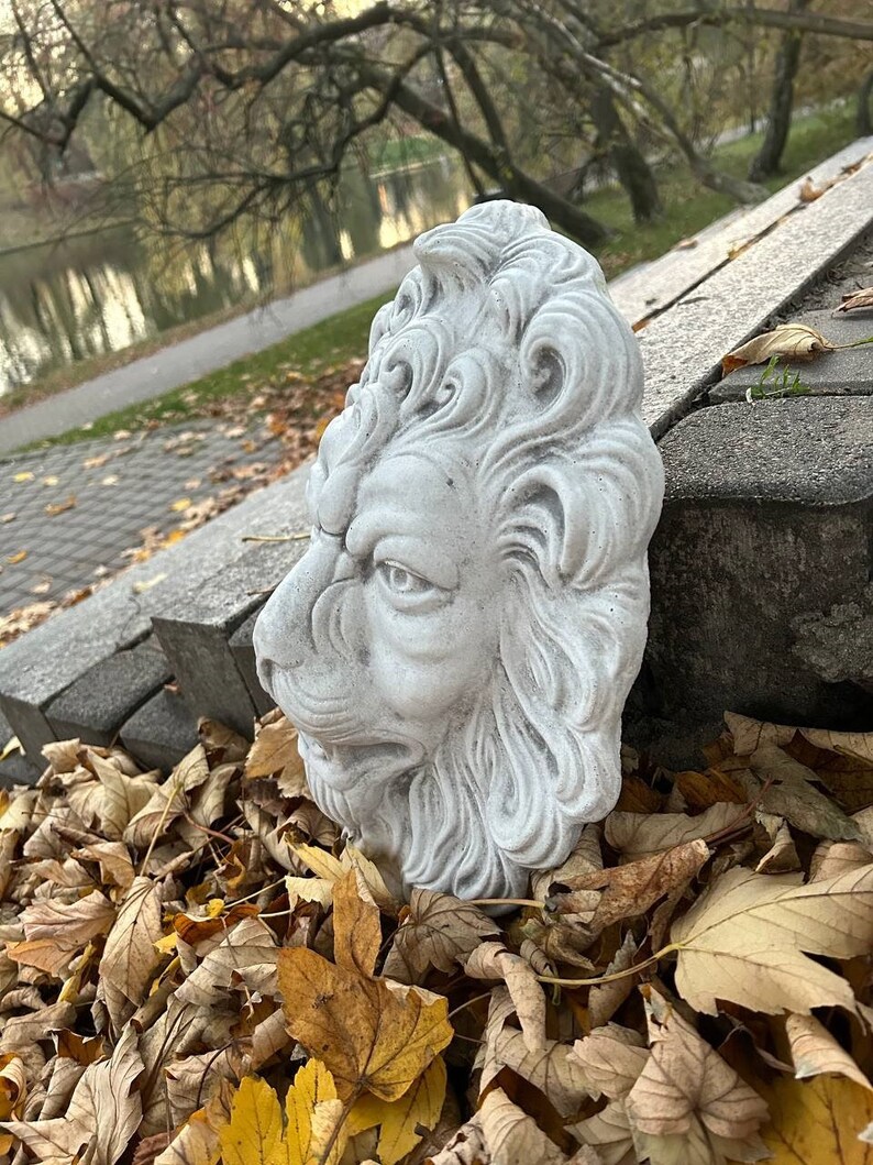 May include: A gray stone lion head sculpture with detailed carvings. The sculpture is placed on a pile of brown leaves.