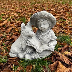 May include: A gray concrete garden statue depicting a young child wearing a straw hat, reading a book with a dog. The statue is set amidst fallen brown leaves and green grass, creating a serene outdoor scene.
