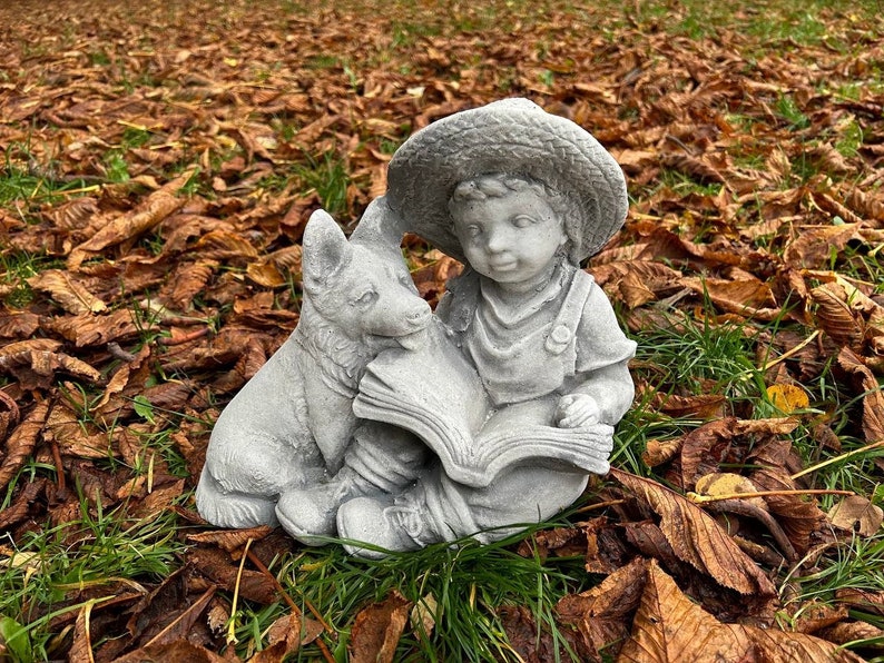 May include: A gray concrete garden statue depicting a young child wearing a straw hat, reading a book with a dog. The statue is set amidst fallen brown leaves and green grass, creating a serene outdoor scene.