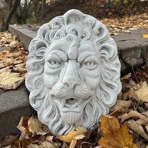 May include: A white concrete lion head sculpture with a detailed mane and a carved mouth. The sculpture is sitting on a bed of fallen autumn leaves.