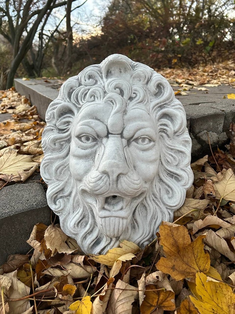 May include: A white concrete lion head sculpture with a detailed mane and a carved mouth. The sculpture is sitting on a bed of fallen autumn leaves.