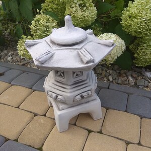 Puede incluir: Una linterna de pagodas de hormig&oacute;n blanco con un dise&ntilde;o decorativo. La linterna est&aacute; sentada en un patio de ladrillo gris y marr&oacute;n.