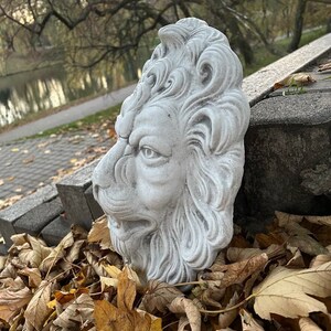 May include: A white concrete lion head sculpture with detailed carvings. The sculpture is sitting on a bed of brown and yellow leaves.