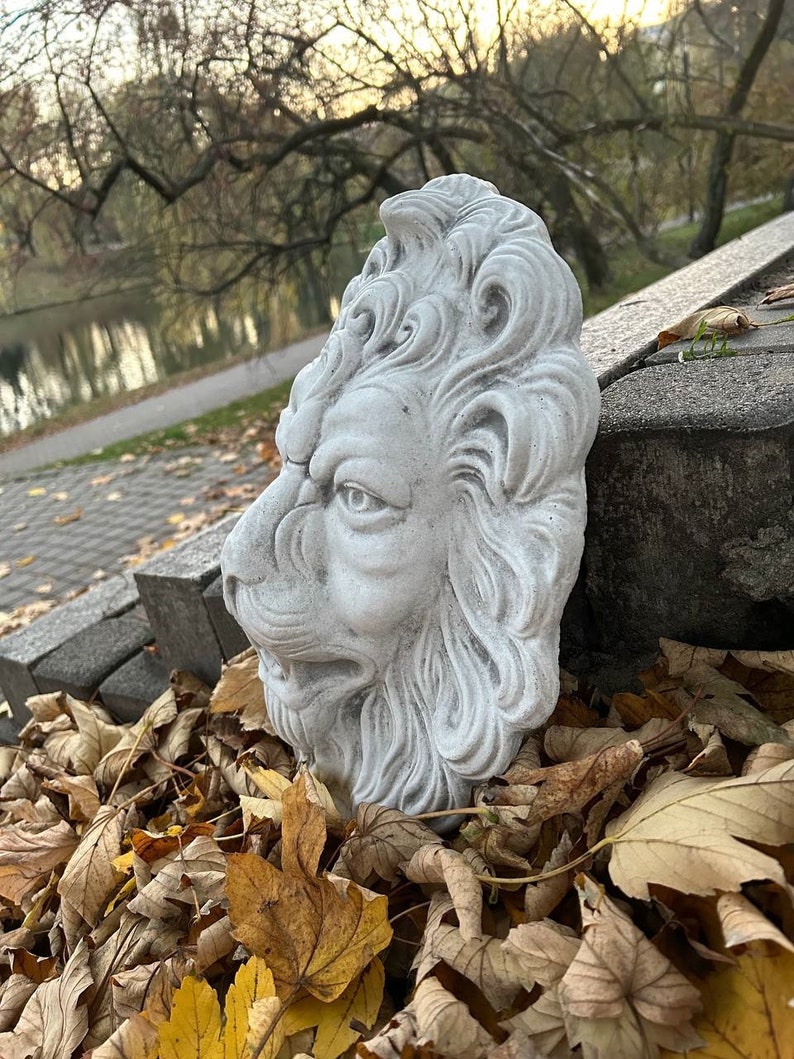 May include: A white concrete lion head sculpture with detailed carvings. The sculpture is sitting on a bed of brown and yellow leaves.