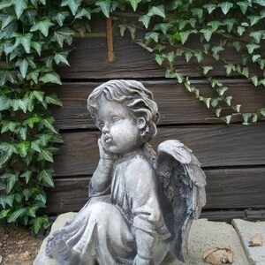 May include: A gray stone sculpture of an angel with wings sitting on a stone base. The angel is looking down and has its hand on its chin. The sculpture is in front of a wooden wall with ivy growing on it.