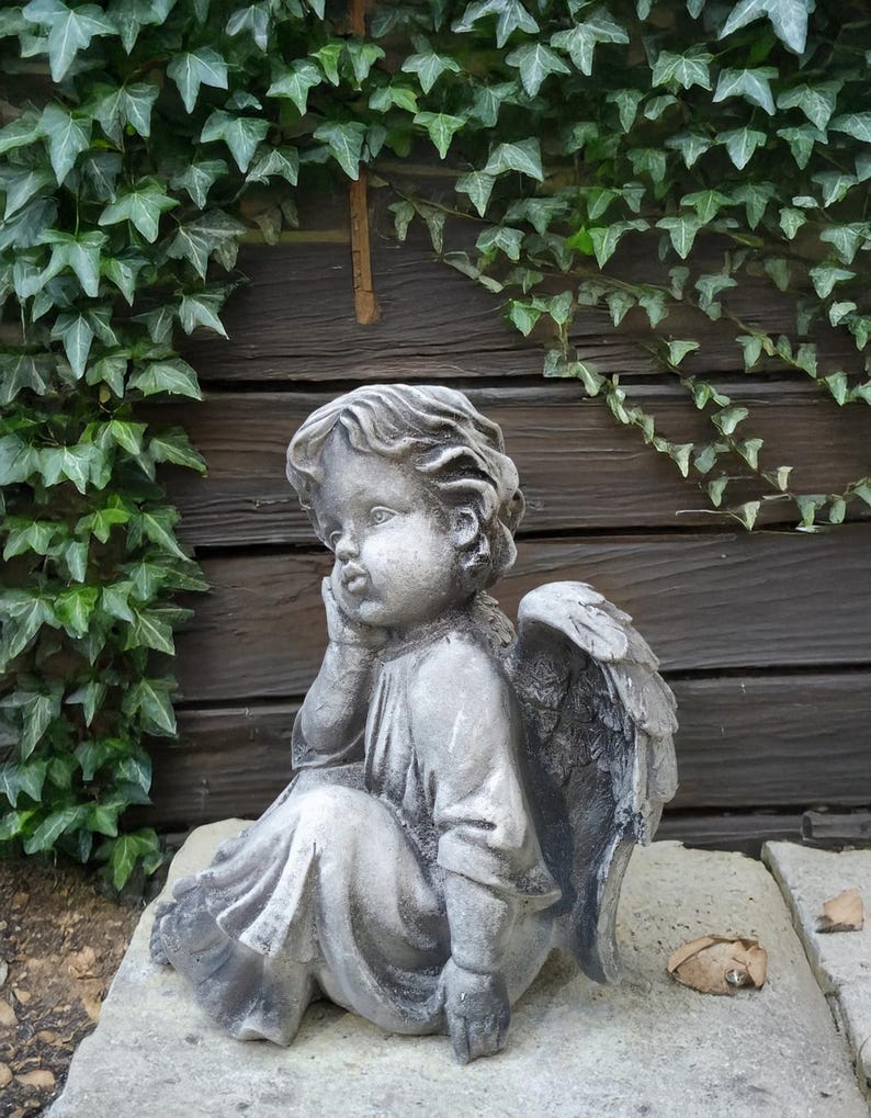 May include: A gray stone sculpture of an angel with wings sitting on a stone base. The angel is looking down and has its hand on its chin. The sculpture is in front of a wooden wall with ivy growing on it.