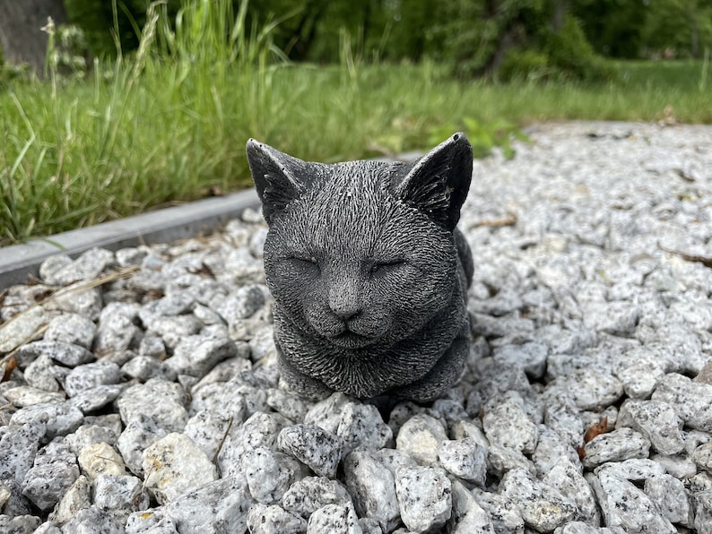 Puede incluir: Una estatua de gato de hormig&oacute;n gris con los ojos cerrados, tumbada en un lecho de peque&ntilde;as piedras grises.