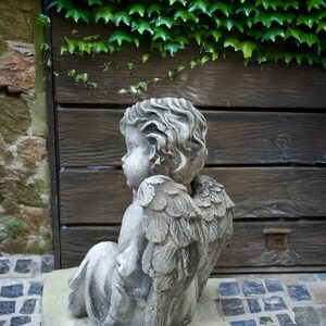 May include: A gray stone cherub statue with wings, sitting on a stone base. The cherub is looking to the left and has a soft, gentle expression.