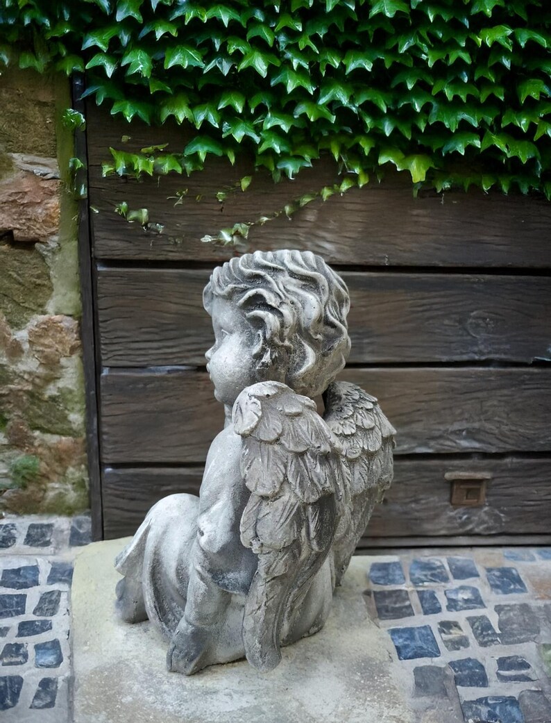 May include: A gray stone cherub statue with wings, sitting on a stone base. The cherub is looking to the left and has a soft, gentle expression.