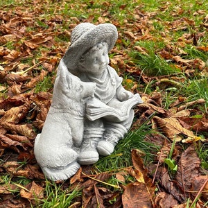 May include: A gray concrete garden statue of a child reading a book, accompanied by a dog. The child wears a hat and is seated, with the dog beside them. The statue is set amidst fallen brown leaves and green grass.