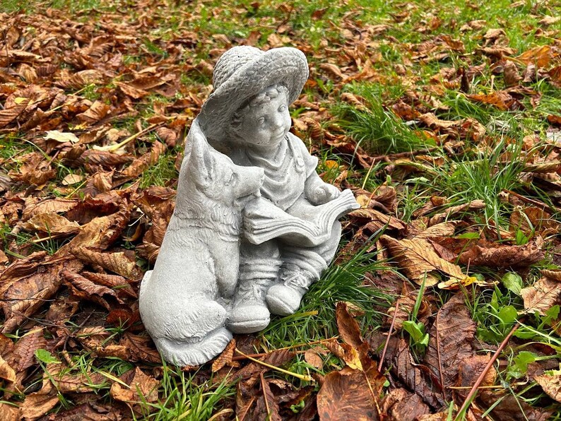 May include: A gray concrete garden statue of a child reading a book, accompanied by a dog. The child wears a hat and is seated, with the dog beside them. The statue is set amidst fallen brown leaves and green grass.