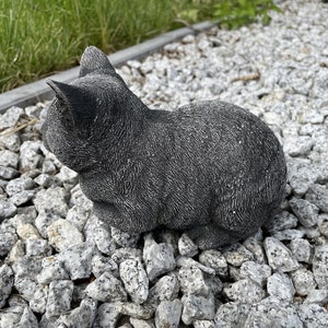 Puede incluir: Una figurita de gato de hormig&oacute;n gris sentada en un lecho de grava blanca.