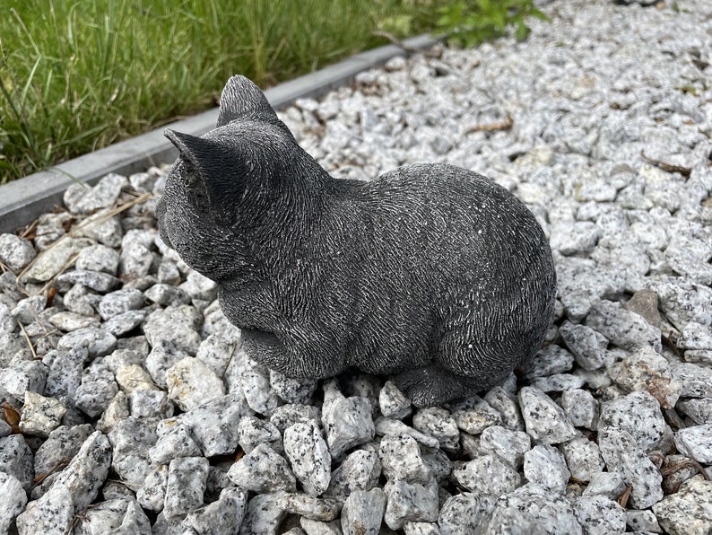 Puede incluir: Una figurita de gato de hormig&oacute;n gris sentada en un lecho de grava blanca.