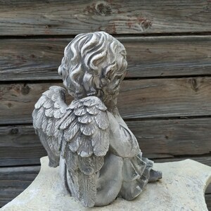 May include: A gray stone cherub statue with wings, sitting on a white stone base. The cherub is looking down and has its wings folded behind it.