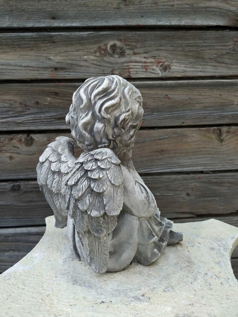 May include: A gray stone cherub statue with wings, sitting on a white stone base. The cherub is looking down and has its wings folded behind it.
