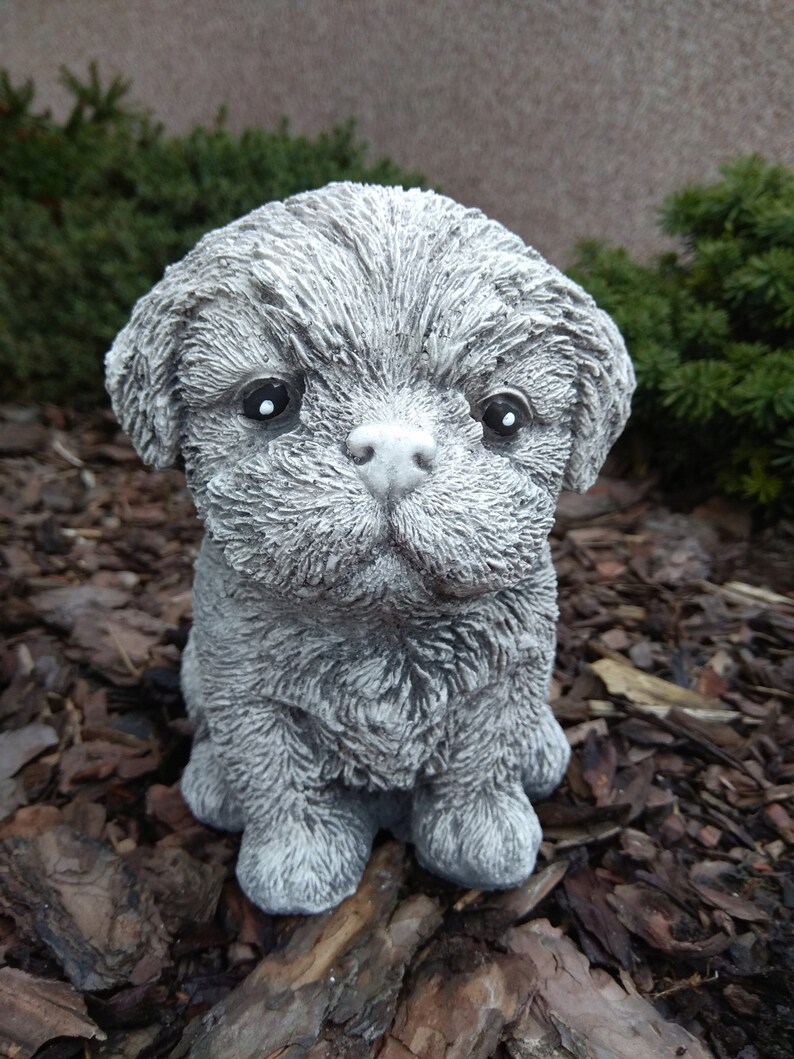 May include: A small, gray, concrete sculpture of a dog with a fluffy texture. The dog is sitting with its paws tucked underneath it and has a sweet expression on its face.