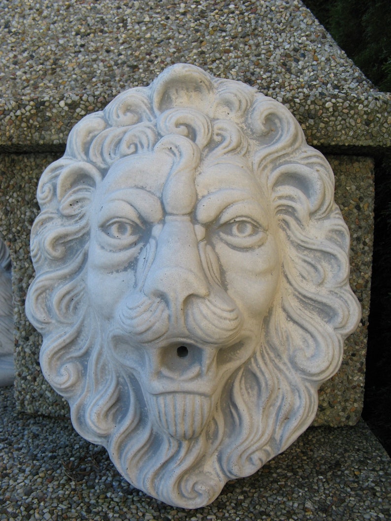 May include: A white stone lion head sculpture with a detailed mane and a carved mouth with a hole for water to flow through. The sculpture is a garden decoration.
