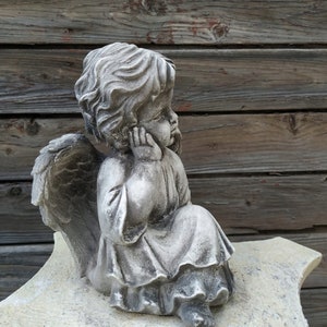 May include: A small, gray stone angel statue with wings, sitting on a light gray stone base. The angel has a thoughtful expression and is holding its chin with its hand.