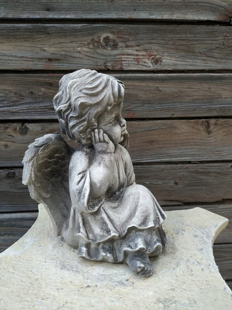 May include: A small, gray stone angel statue with wings, sitting on a light gray stone base. The angel has a thoughtful expression and is holding its chin with its hand.