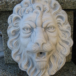 May include: A white concrete lion head sculpture with a detailed mane and a spout in the mouth. The sculpture is a garden decoration.