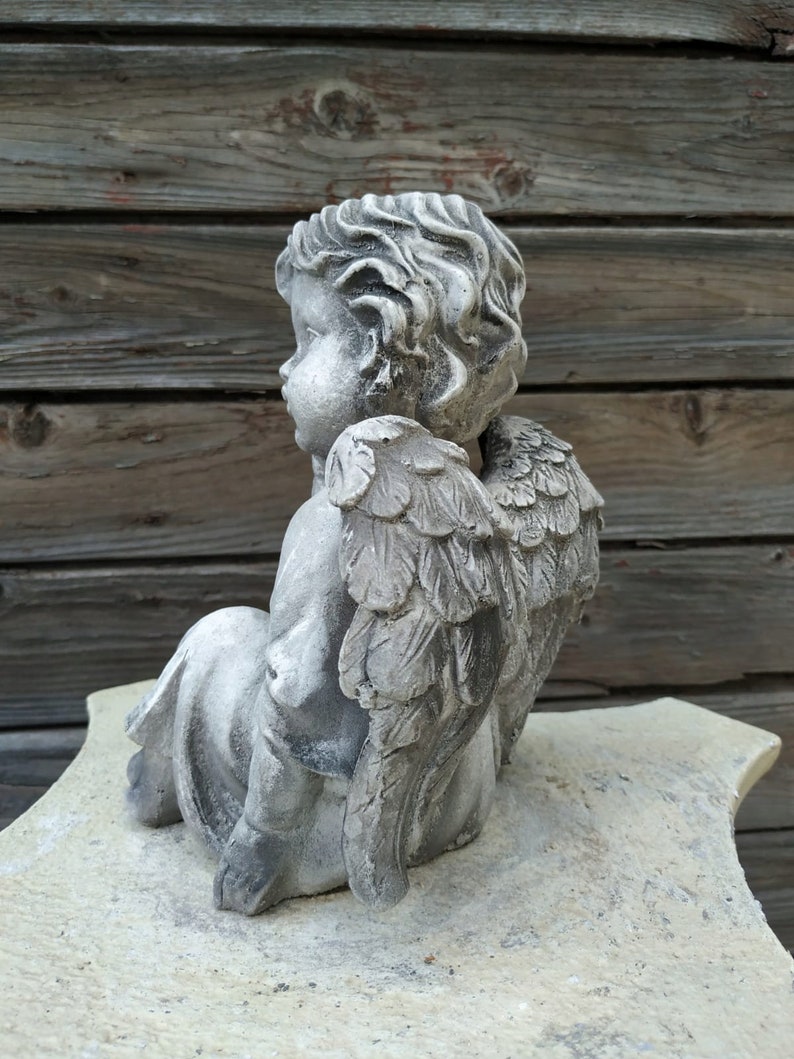 May include: A gray stone cherub statue with wings, sitting on a white stone base. The cherub has a detailed face and curly hair.