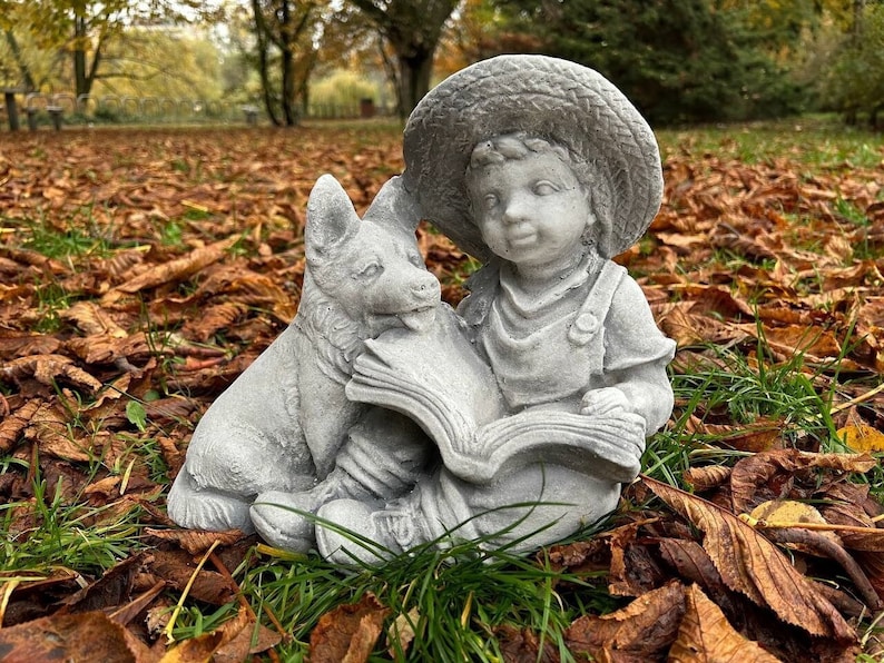 May include: A gray concrete statue of a child wearing overalls and a straw hat, reading a book with a dog by their side. The statue is set outdoors on a bed of fallen leaves and green grass, with a blurred background of trees.