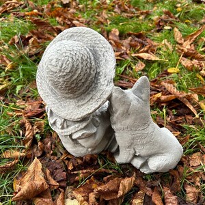 May include: A concrete garden statue of a child wearing a hat, with a dog. The statue is gray and has a weathered appearance. The dog is sitting next to the child, and the child is wearing a hat. The statue is surrounded by grass and leaves.