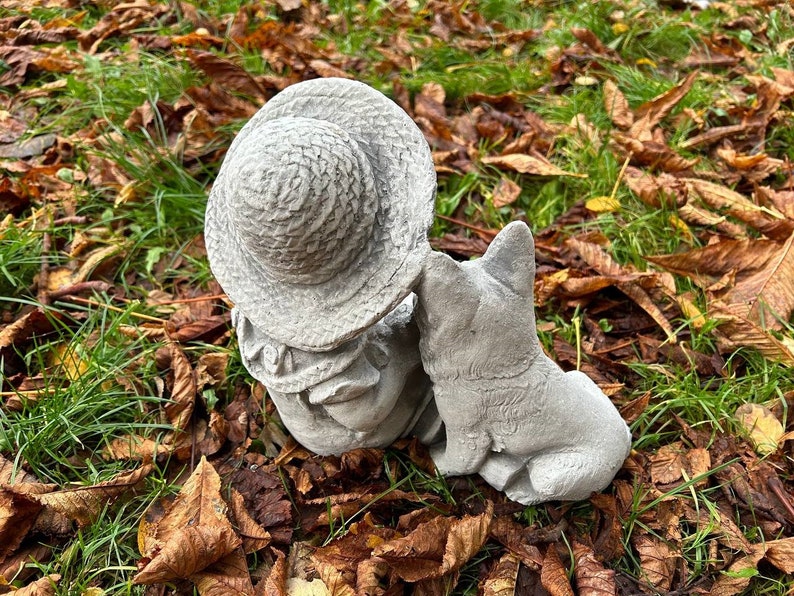 May include: A concrete garden statue of a child wearing a hat, with a dog. The statue is gray and has a weathered appearance. The dog is sitting next to the child, and the child is wearing a hat. The statue is surrounded by grass and leaves.