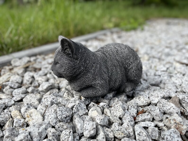 Puede incluir: Una escultura de gato de hormig&oacute;n gris en una pose de dormir. La escultura est&aacute; sentada en un lecho de peque&ntilde;as piedras grises.