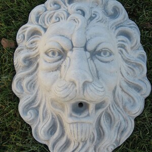 May include: A gray stone lion head sculpture with detailed carvings. The sculpture has a hole in the mouth, suggesting it may be a fountain or water feature.