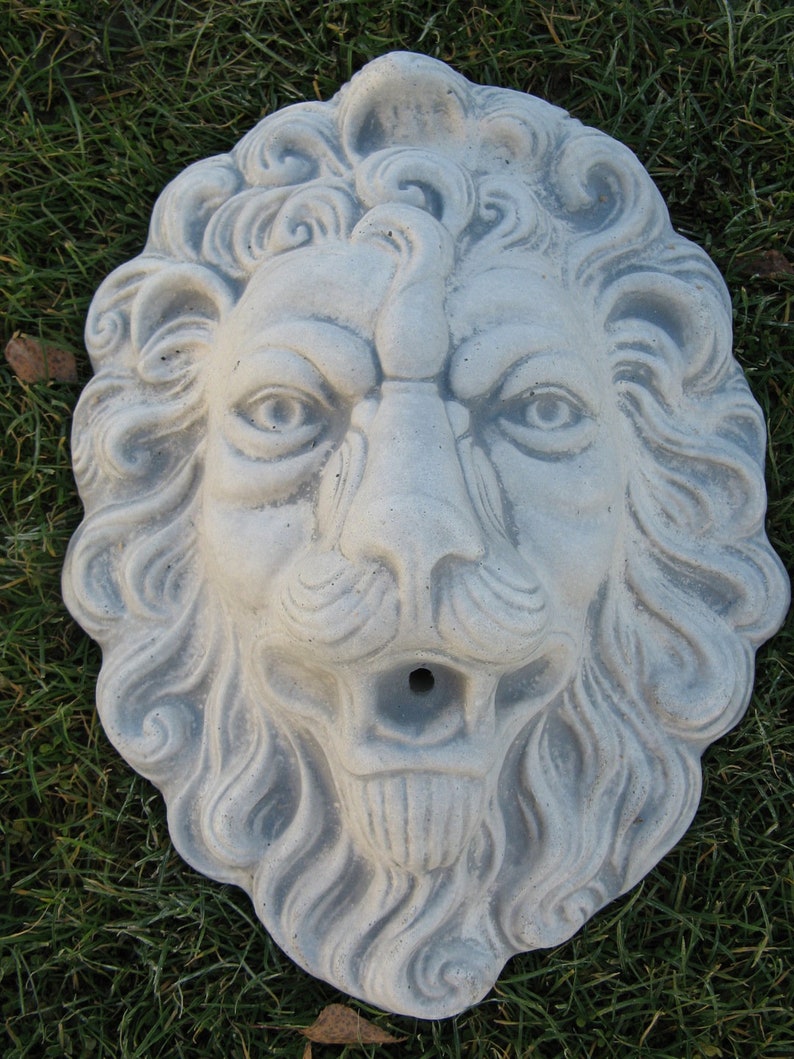 May include: A gray stone lion head sculpture with detailed carvings. The sculpture has a hole in the mouth, suggesting it may be a fountain or water feature.