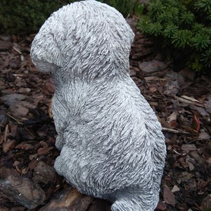 May include: A gray concrete sculpture of a small dog sitting with its head down. The sculpture is detailed with fur texture and is sitting on a bed of brown wood chips.