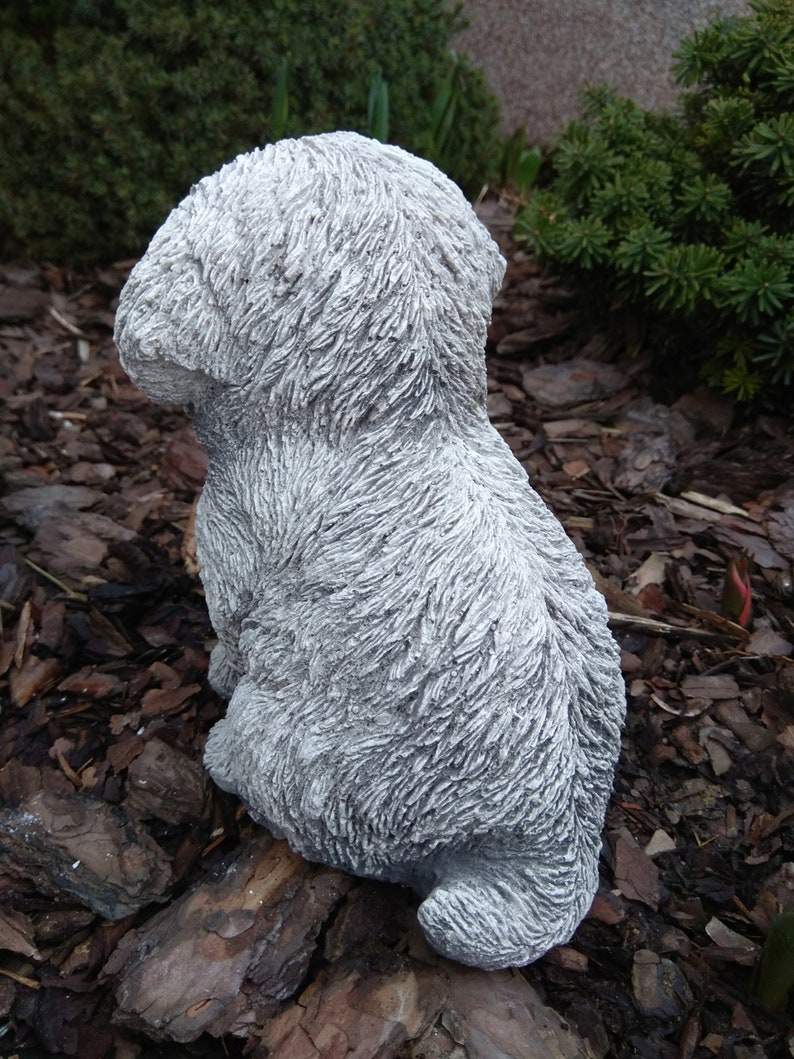 May include: A gray concrete sculpture of a small dog sitting with its head down. The sculpture is detailed with fur texture and is sitting on a bed of brown wood chips.