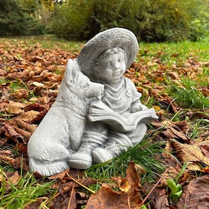 May include: A gray concrete garden statue depicts a young child wearing a hat, reading a book, with a dog companion. The statue is set in a natural outdoor environment with fallen leaves and green grass.