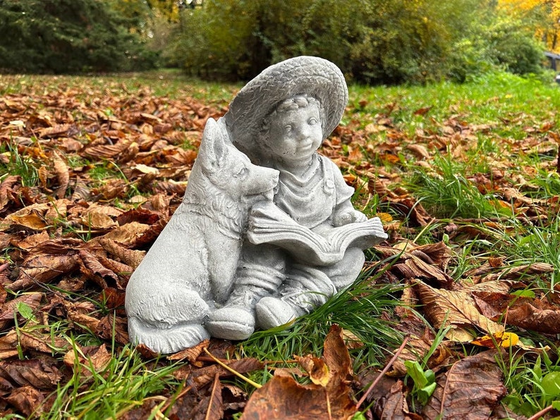 May include: A gray concrete garden statue depicts a young child wearing a hat, reading a book, with a dog companion. The statue is set in a natural outdoor environment with fallen leaves and green grass.