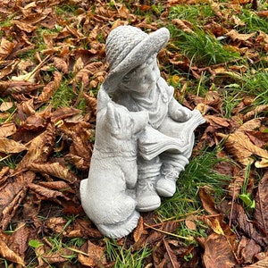 May include: A concrete garden statue depicting a child in a hat reading a book, accompanied by a dog. The statue is gray and detailed, set amidst fallen brown leaves and green grass.