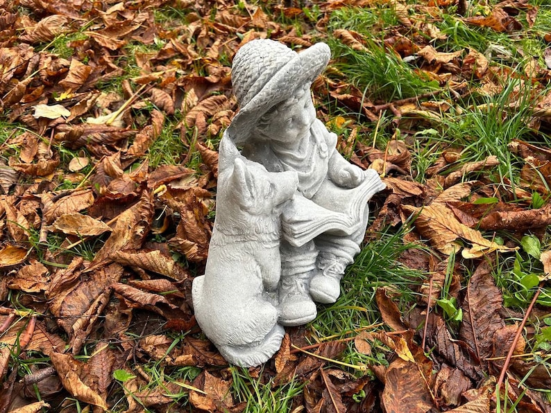 May include: A concrete garden statue depicting a child in a hat reading a book, accompanied by a dog. The statue is gray and detailed, set amidst fallen brown leaves and green grass.