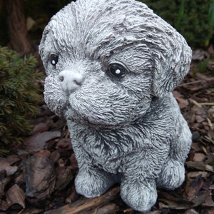 May include: A small, gray, furry, concrete statue of a dog sitting on a bed of wood chips.