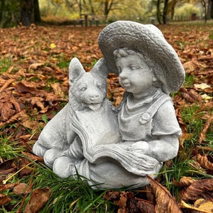 May include: A gray concrete garden statue of a young child wearing a straw hat, reading a book with a dog. The statue is set in a bed of fallen leaves and grass, with a blurred park background.