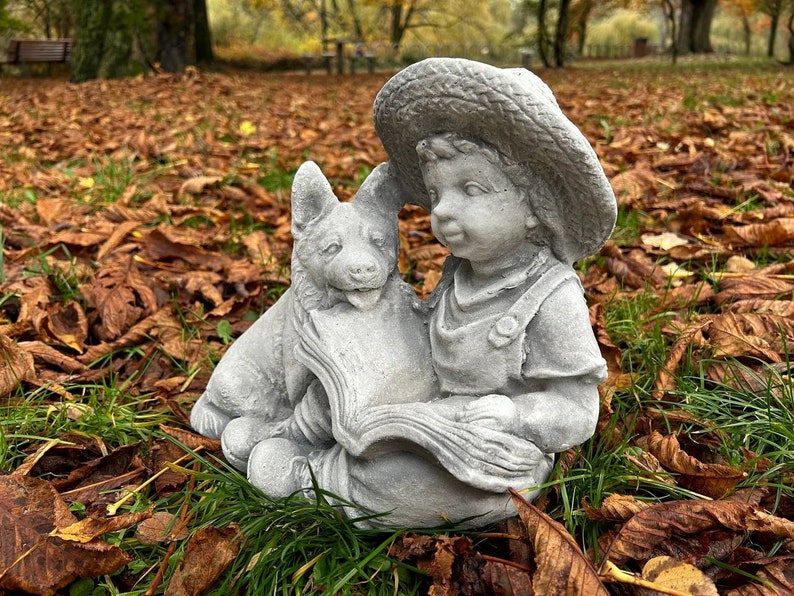 May include: A gray concrete garden statue of a young child wearing a straw hat, reading a book with a dog. The statue is set in a bed of fallen leaves and grass, with a blurred park background.