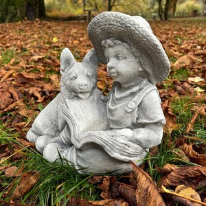 May include: A gray concrete garden statue depicting a young child wearing a straw hat and overalls, reading a book with a dog. The statue is set in a bed of fallen leaves and grass, creating a serene outdoor scene.