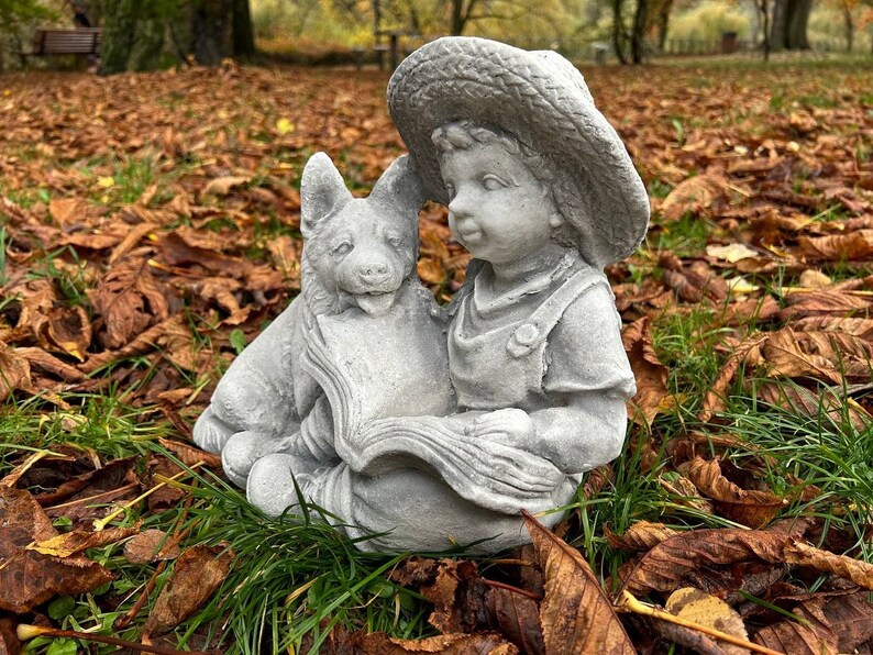 May include: A gray concrete garden statue depicting a young child wearing a straw hat and overalls, reading a book with a dog. The statue is set in a bed of fallen leaves and grass, creating a serene outdoor scene.