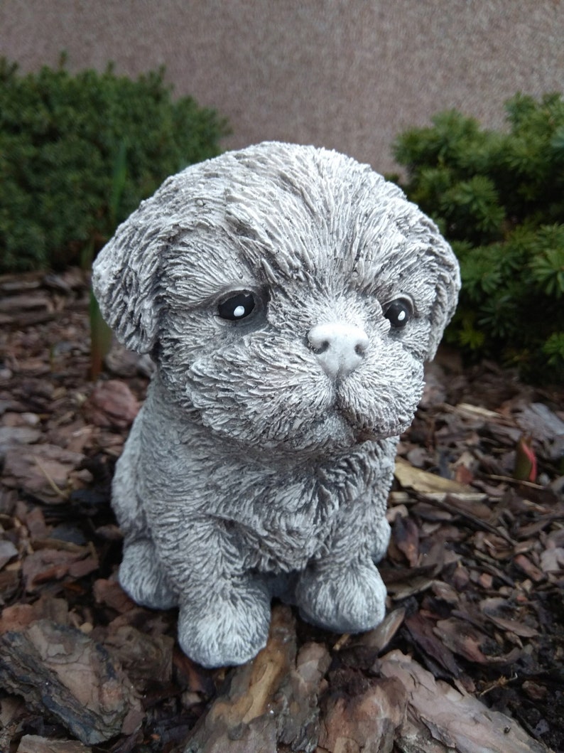 May include: A small, gray, concrete statue of a dog with a fluffy, textured coat. The dog is sitting with its head tilted down and its eyes closed.