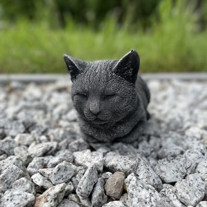 Puede incluir: Una figurita de gato gris durmiendo hecha de piedra o resina. El gato est&aacute; enroscado con los ojos cerrados y descansa sobre una cama de peque&ntilde;as piedras grises.