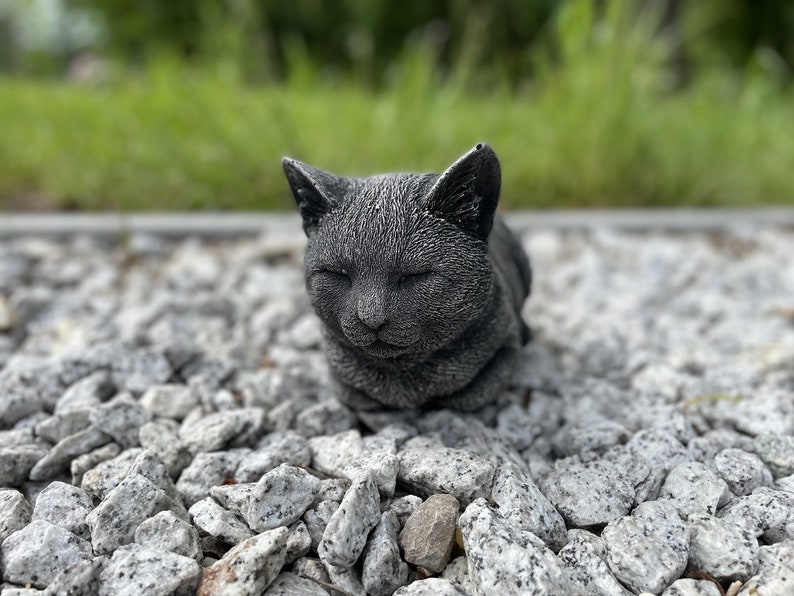 Puede incluir: Una figurita de gato gris durmiendo hecha de piedra o resina. El gato est&aacute; enroscado con los ojos cerrados y descansa sobre una cama de peque&ntilde;as piedras grises.