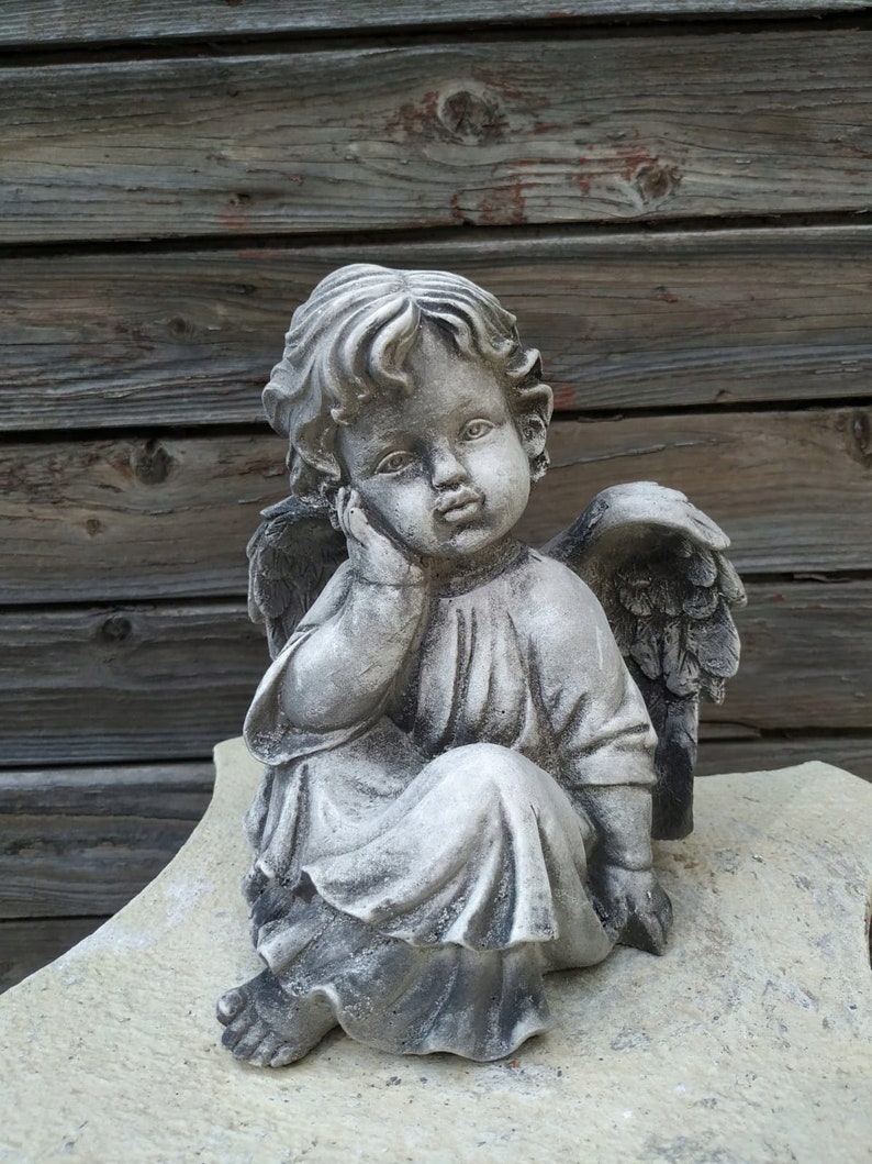 May include: A gray stone cherub statue with wings, sitting with its head resting on its hand. The cherub is wearing a flowing robe and has a sweet expression on its face.