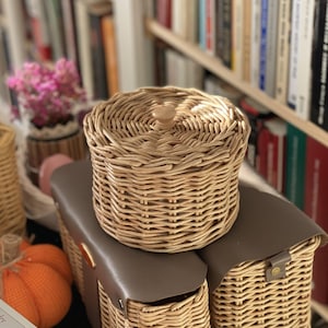 May include: A round, light-tan woven wicker basket with a matching lid featuring a small central knob. The basket sits atop two rectangular woven wicker containers, each topped with a smooth, dark-brown faux leather flap and a small metal fastener.