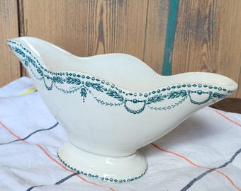 May include: A white ceramic gravy boat with a scalloped rim and pedestal base. The boat is decorated with a teal floral and beaded design around the rim. It sits on a striped cloth with a wooden spoon.