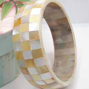 Peut inclure: Un large bracelet rond avec un motif en damier de carrés blancs et jaunes. Le bracelet est fait d'un matériau brillant et a une surface lisse et incurvée. Le bracelet mesure environ 7.6 cm de large.