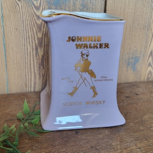 Op de afbeelding: Een paarse keramische kan met een gouden rand en een Johnnie Walker-logo. Het logo toont een man in een wandelende pose met de tekst "Born 1820 - Still Going Strong" en "Scotch Whisky".